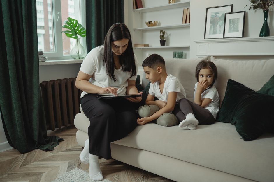 Mother and children sitting together on a sofa, engaged with a digital tablet indoors.