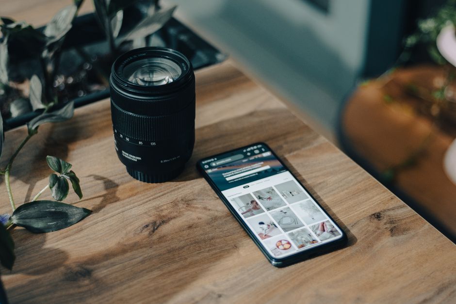 A smartphone and camera lens placed on a wooden table surrounded by plants for a creative photography setup.